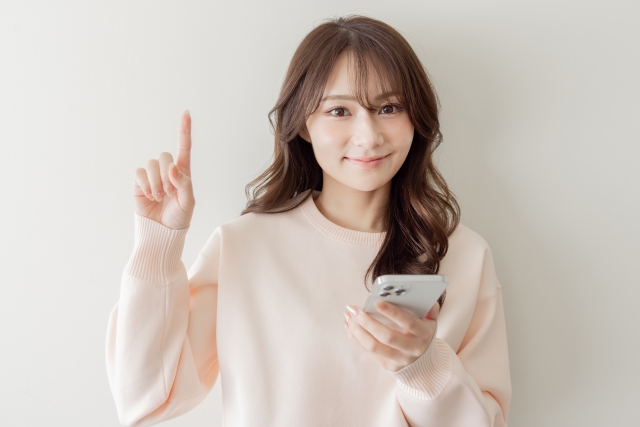 スマホを手に持ち指を立てる仕草をする女性の画像