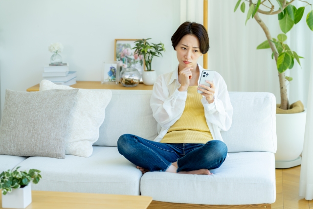 ソファに座ってスマホを見ながら悩んだ表情をしている女性の画像