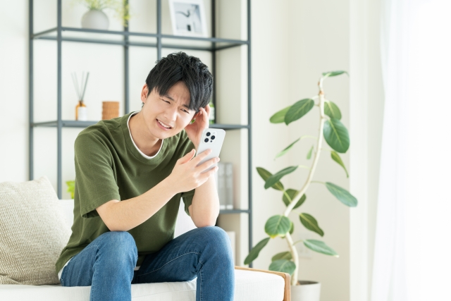 ソファに座って悩んだ表情でスマホを見ている男性